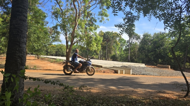 Aurovilietis važiuoja motociklu pro per mišką statomą kelią